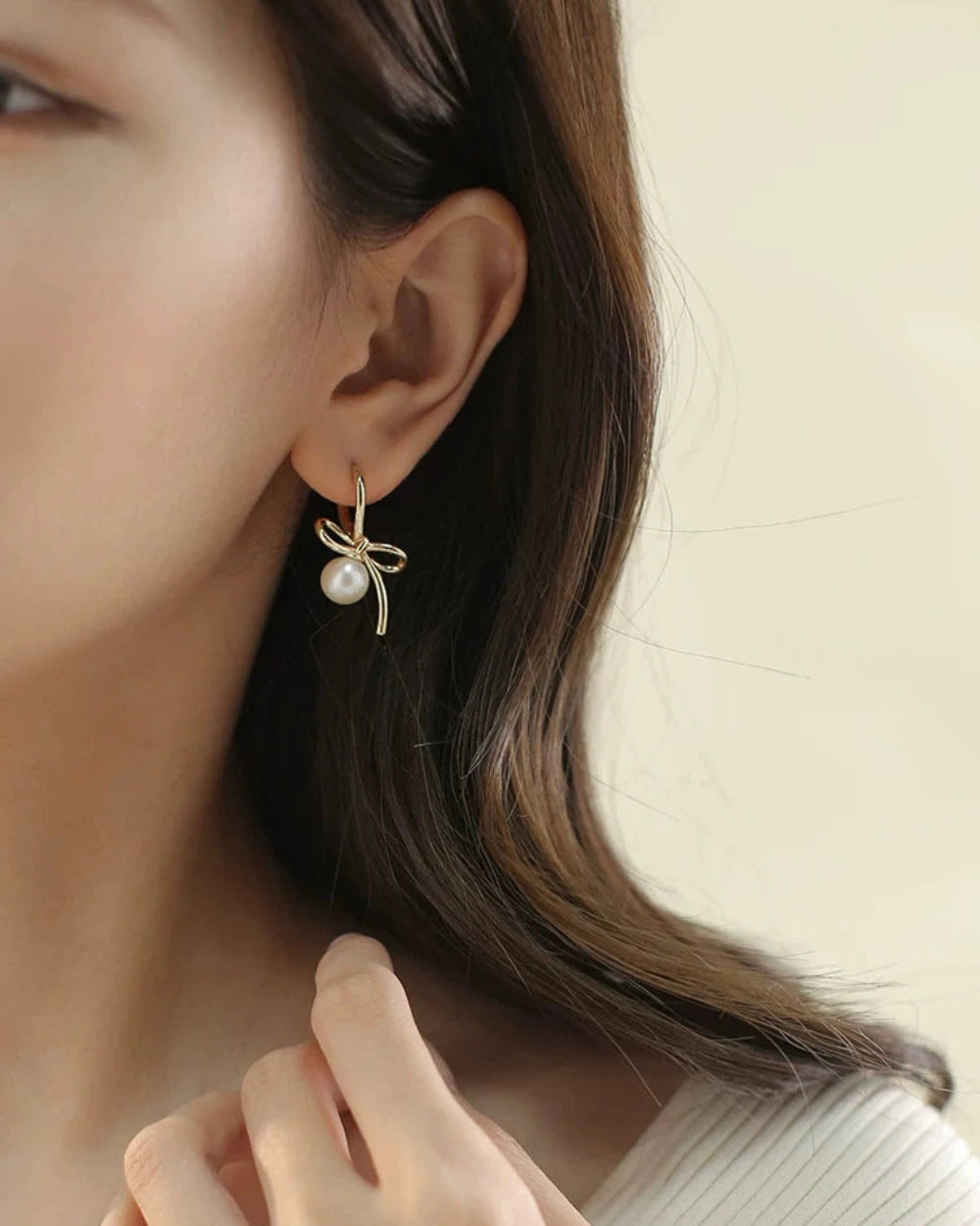 Close-up of a person wearing a gold earring with a pearl on a neutral background