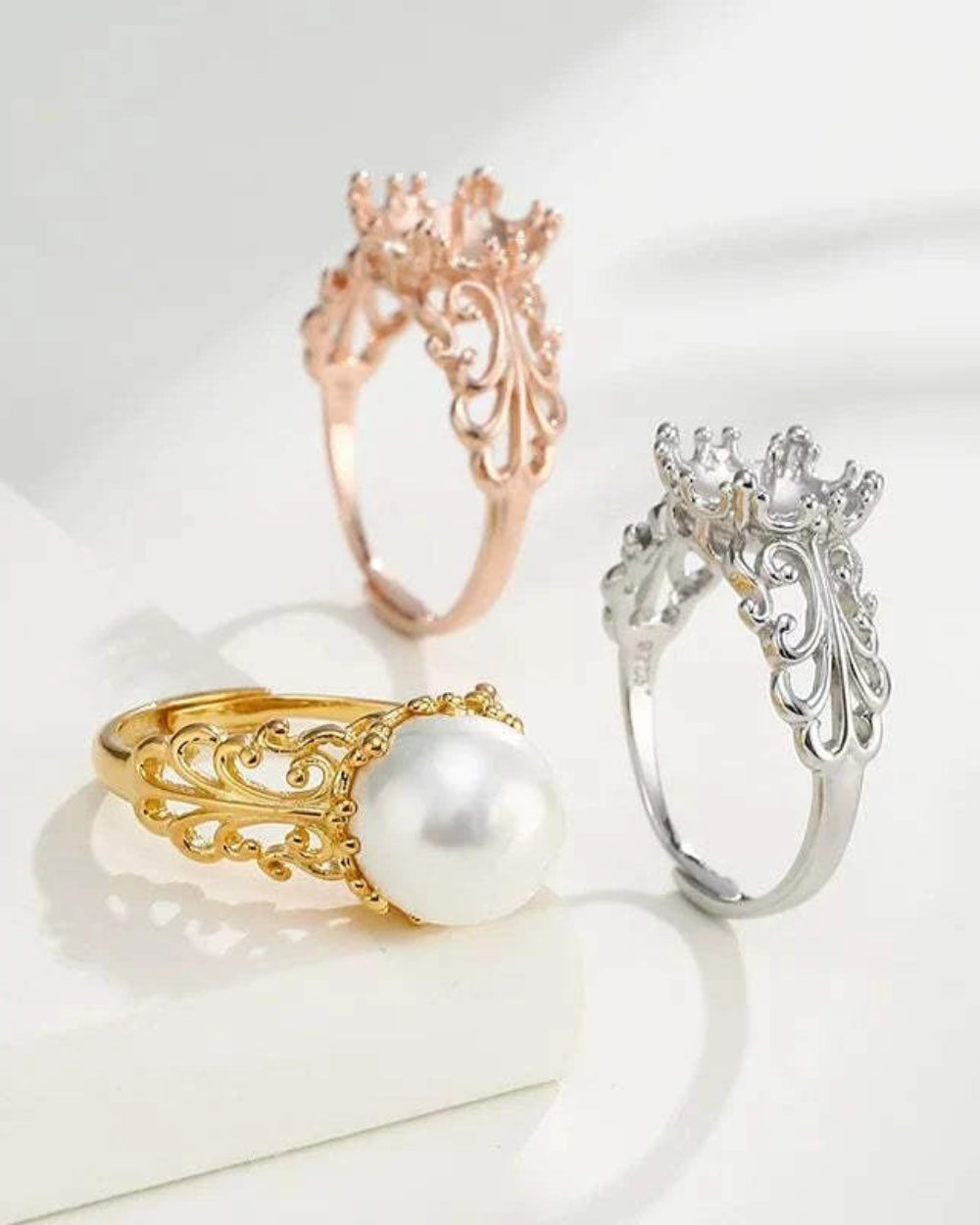 Three ornate rings in gold, rose gold, and silver with pearls on a light background
