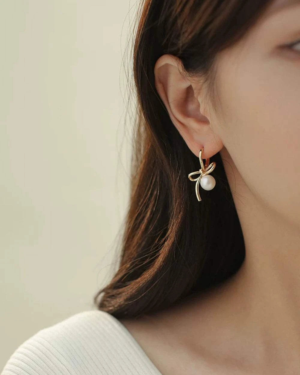 Close-up of a person wearing a gold earring with a pearl on a neutral background