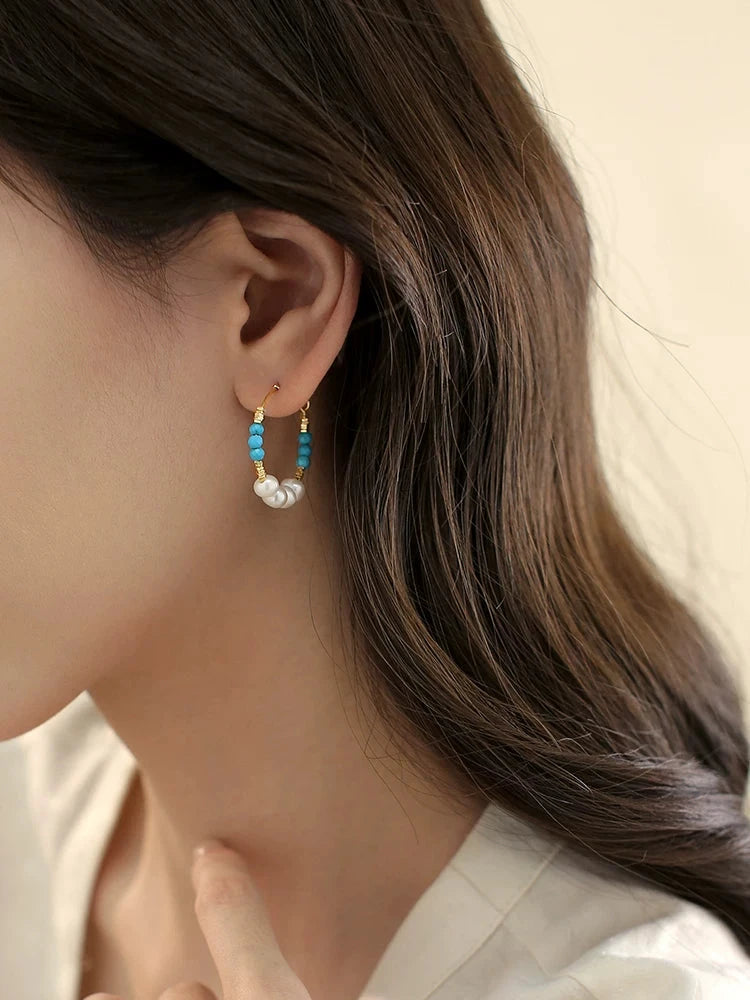 Close-up of a person wearing pearl and turquoise hoop earrings with a neutral background.