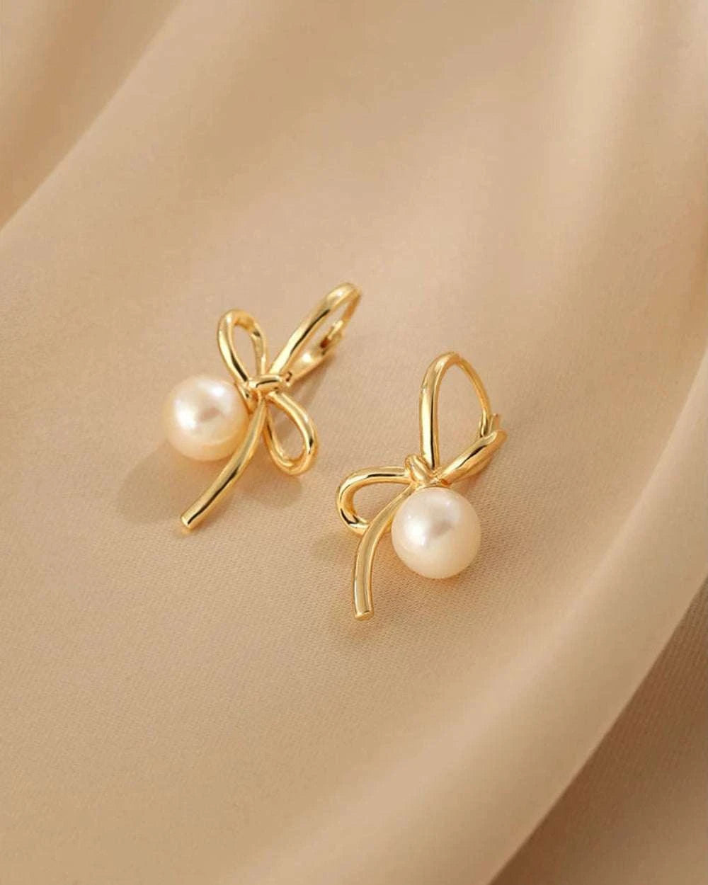Gold bow-shaped earrings with pearl centers on a beige background