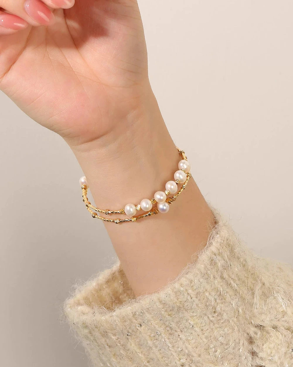 Hand wearing a gold bracelet with pearl accents on a neutral background
