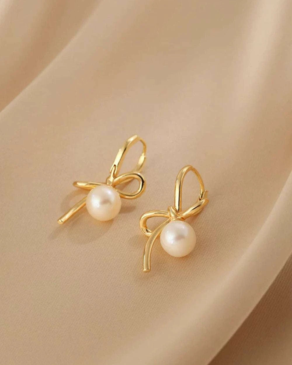 Gold bow-shaped earrings with pearl centers on a beige fabric background